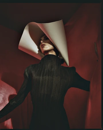 Woman in sculptural white hat and black dress poses against a red backdrop in a studio setting, emphasizing fashion and artistic design.