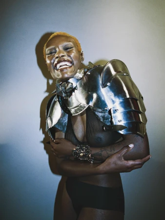 Smiling woman with short blonde hair wearing silver metal shoulder armor and black bikini bottom posing against a light wall with a shadow.