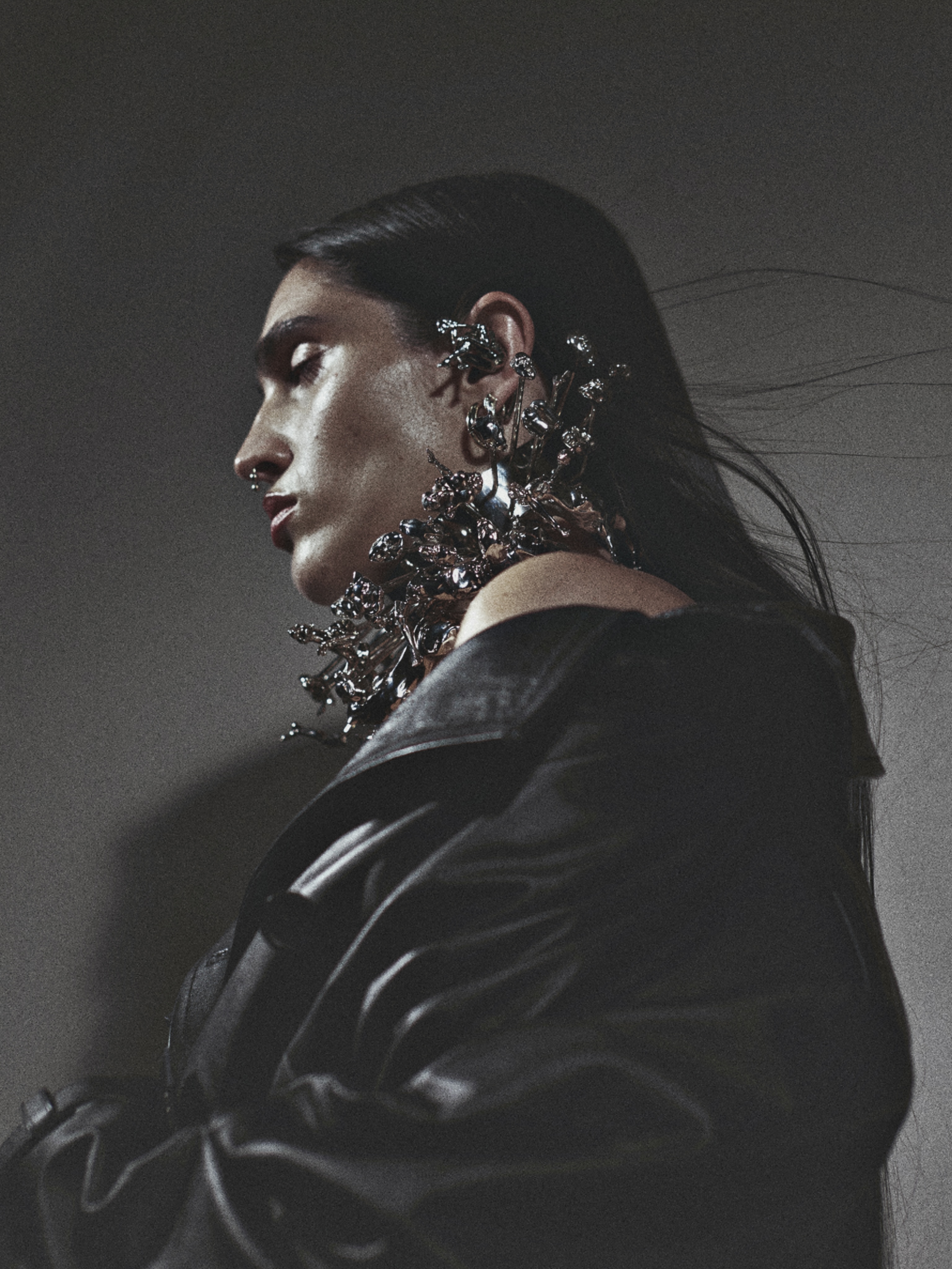 Woman in profile wearing elaborate metallic floral jewelry and a black leather jacket against a grey studio backdrop in a fashion editorial.