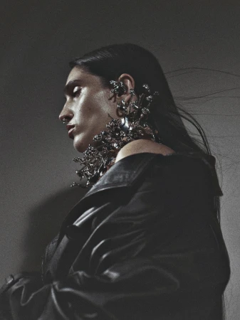 Woman in profile wearing elaborate metallic floral jewelry and a black leather jacket against a grey studio backdrop in a fashion editorial.