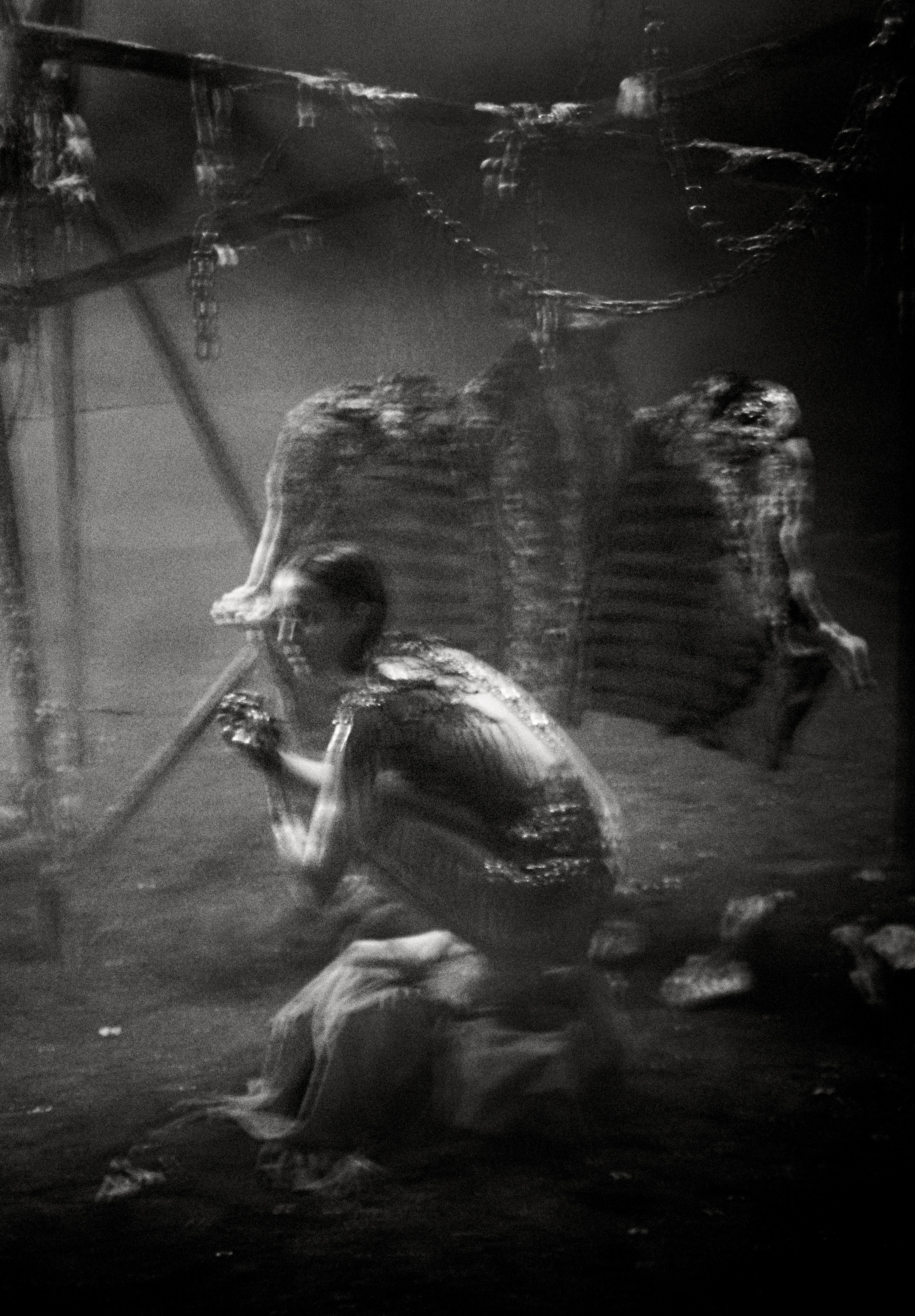 Monochrome image features a woman in a crouched position with ribs hanging in the background, creating a stark and evocative scene.