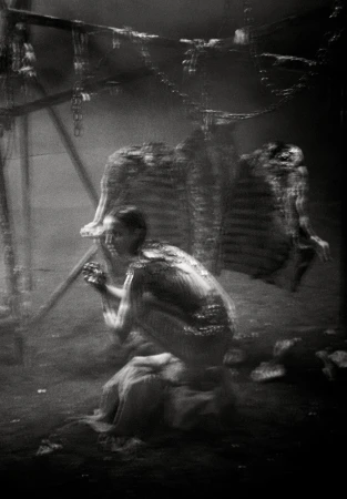 Monochrome image features a woman in a crouched position with ribs hanging in the background, creating a stark and evocative scene.