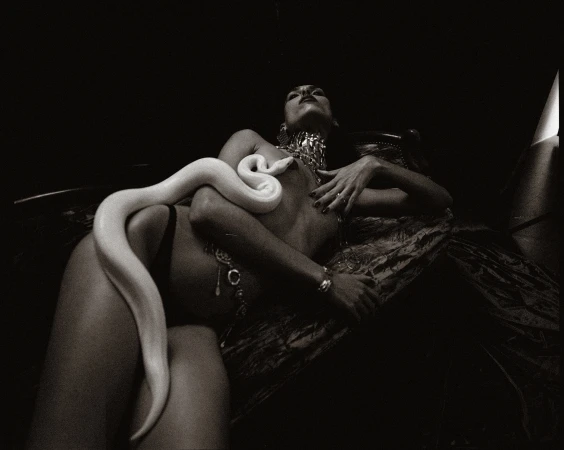 Black and white studio shot of an adult woman lying down with a large white snake, jewelry, and a patterned silk textile. 