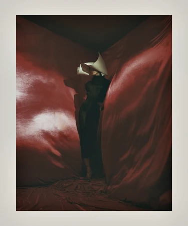 A woman in a black dress and a large white paper hat stands draped in red fabric in a dark room, creating a striking fashion image.