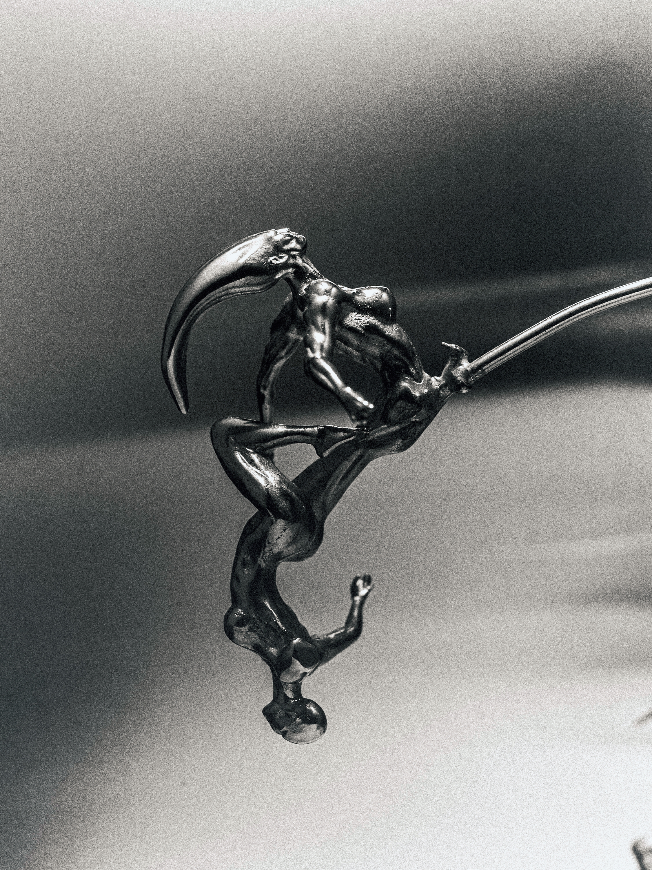 Monochrome photograph of a metallic sculpture depicting two figures in a stylized embrace on a grey gradient background with dramatic lighting.