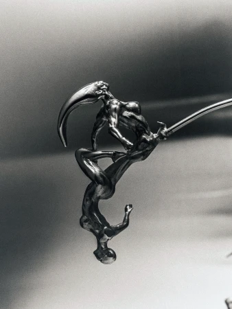 Monochrome photograph of a metallic sculpture depicting two figures in a stylized embrace on a grey gradient background with dramatic lighting.