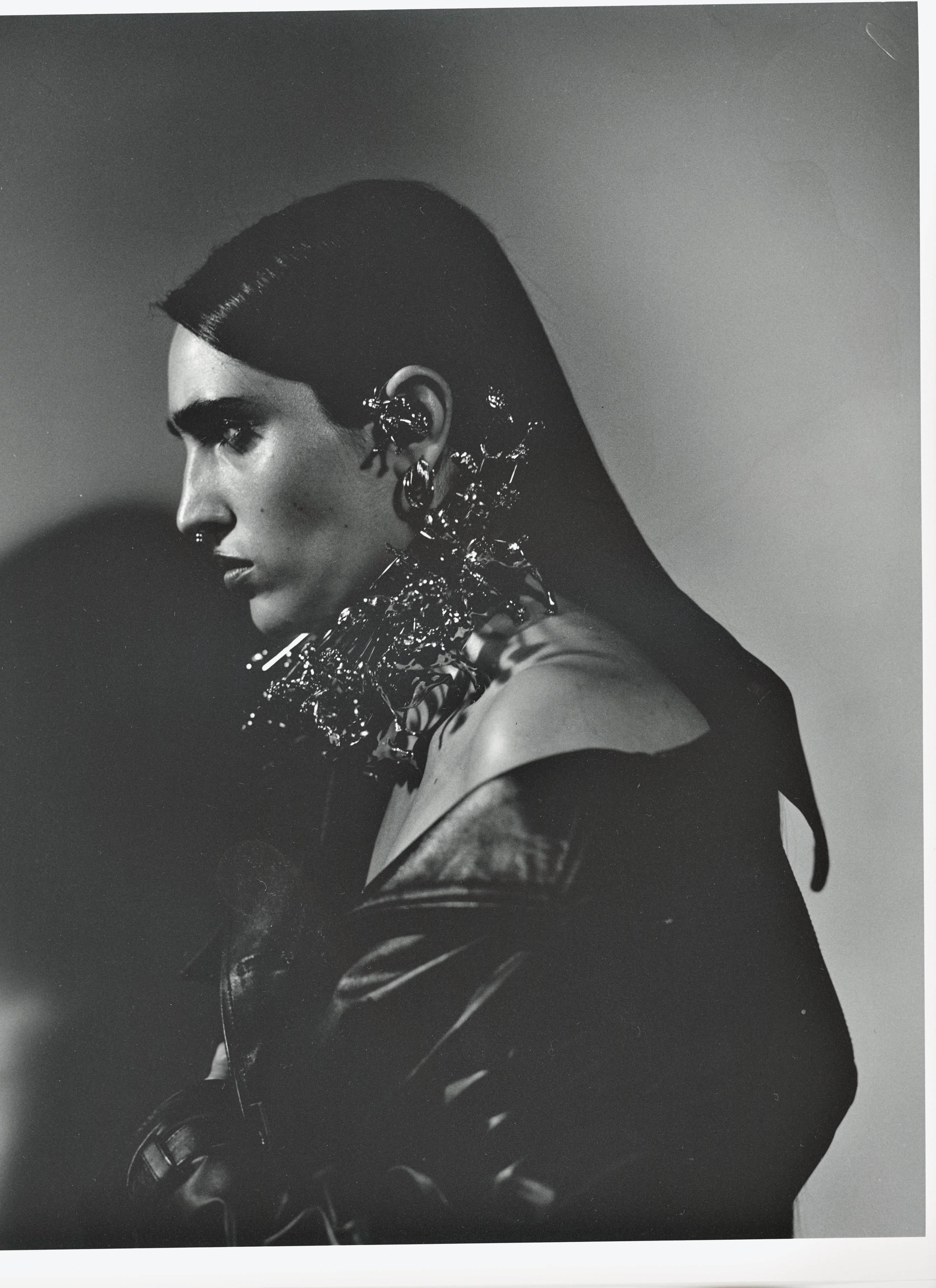 Monochrome portrait of a woman with dark hair, wearing an off-the-shoulder top and striking metal jewelry. Dramatic lighting.