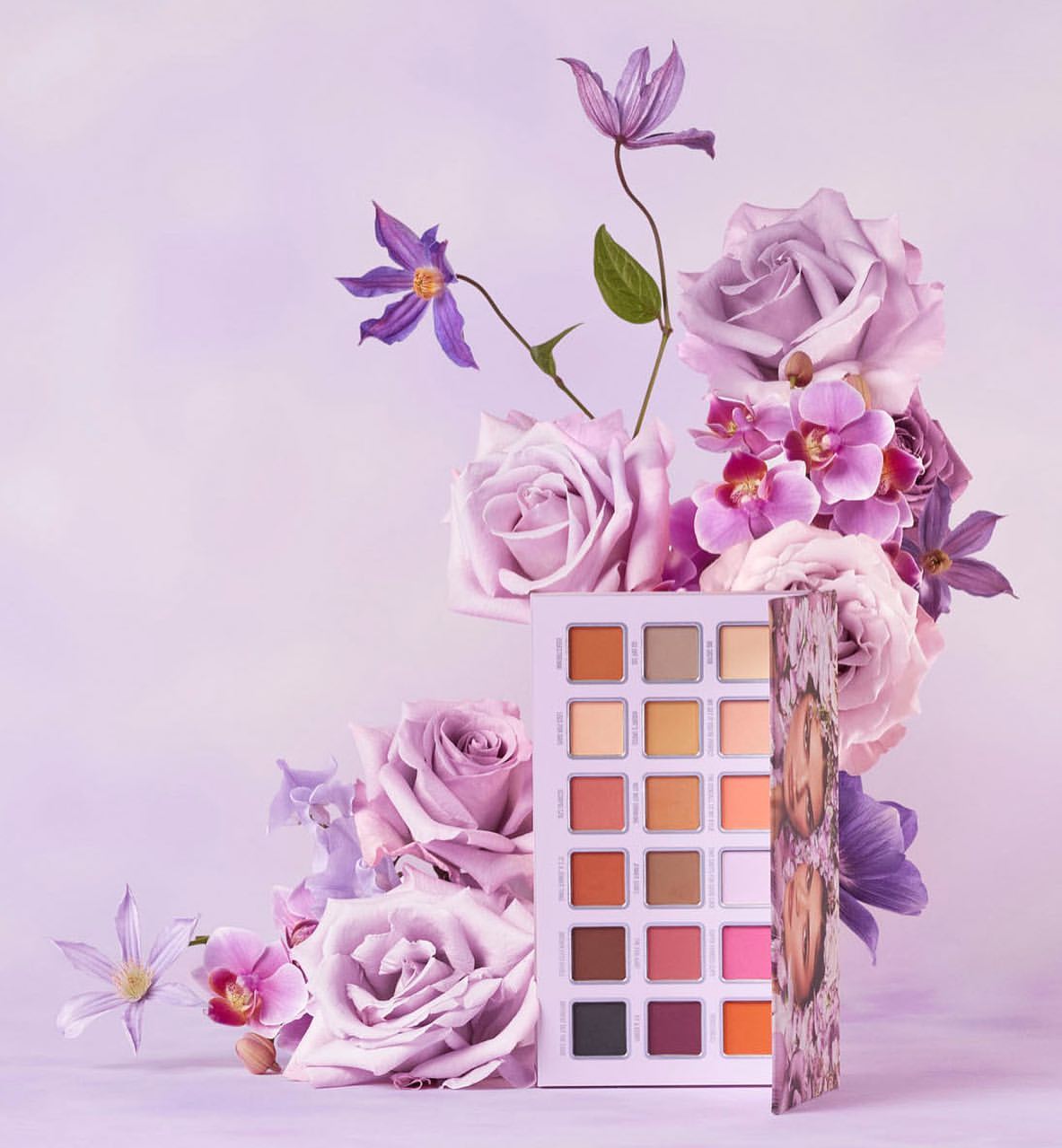 Makeup palette surrounded by purple roses and orchids in a beauty product still life composition against a soft lavender background.
