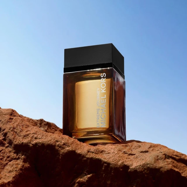 Michael Kors Extreme Journey fragrance bottle on a rock against a clear blue sky, product shot, outdoor setting.