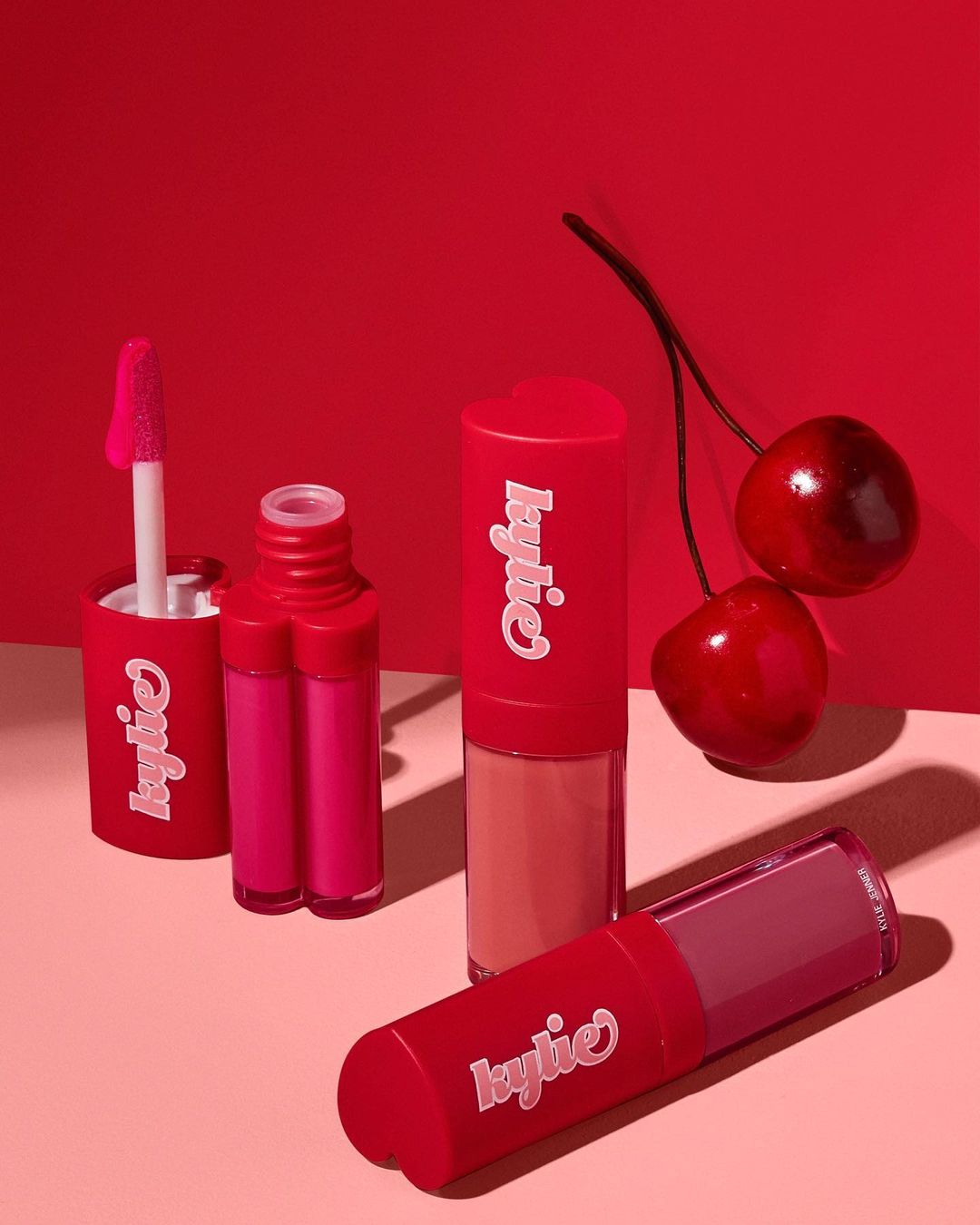 Kylie Cosmetics lip products in red packaging on a pink surface with red cherries, set against a bold red background for beauty advertisement.