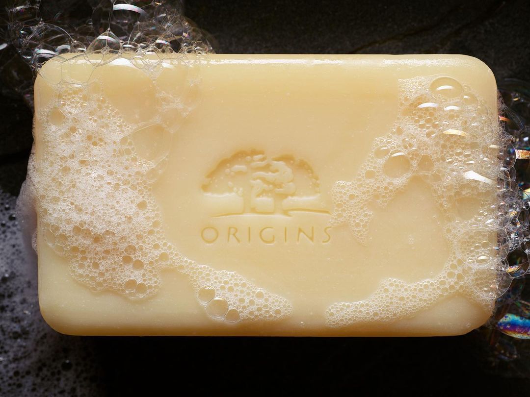 Closeup of a bar of Origins soap covered with white bubbles. The brand's logo of a tree is etched on the surface of the bar.
