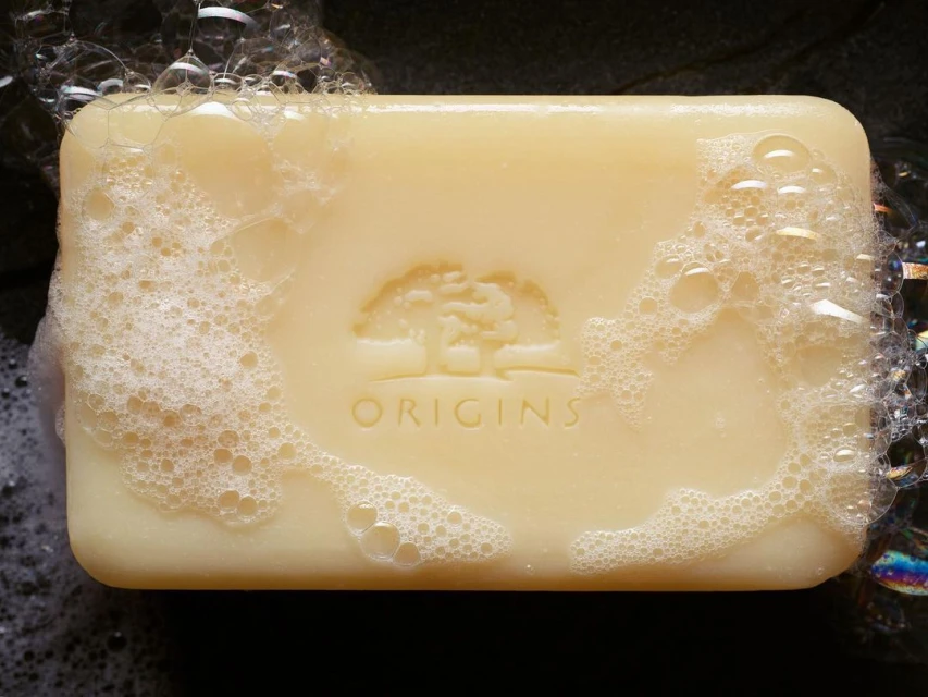 Closeup of a bar of Origins soap covered with white bubbles. The brand's logo of a tree is etched on the surface of the bar.