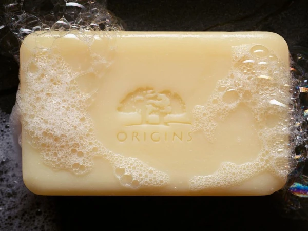 Closeup of a bar of Origins soap covered with white bubbles. The brand's logo of a tree is etched on the surface of the bar.