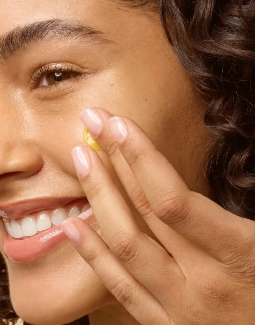 Close-up of a woman applying a round, yellow skincare patch to her cheek, showcasing natural light and skincare application.