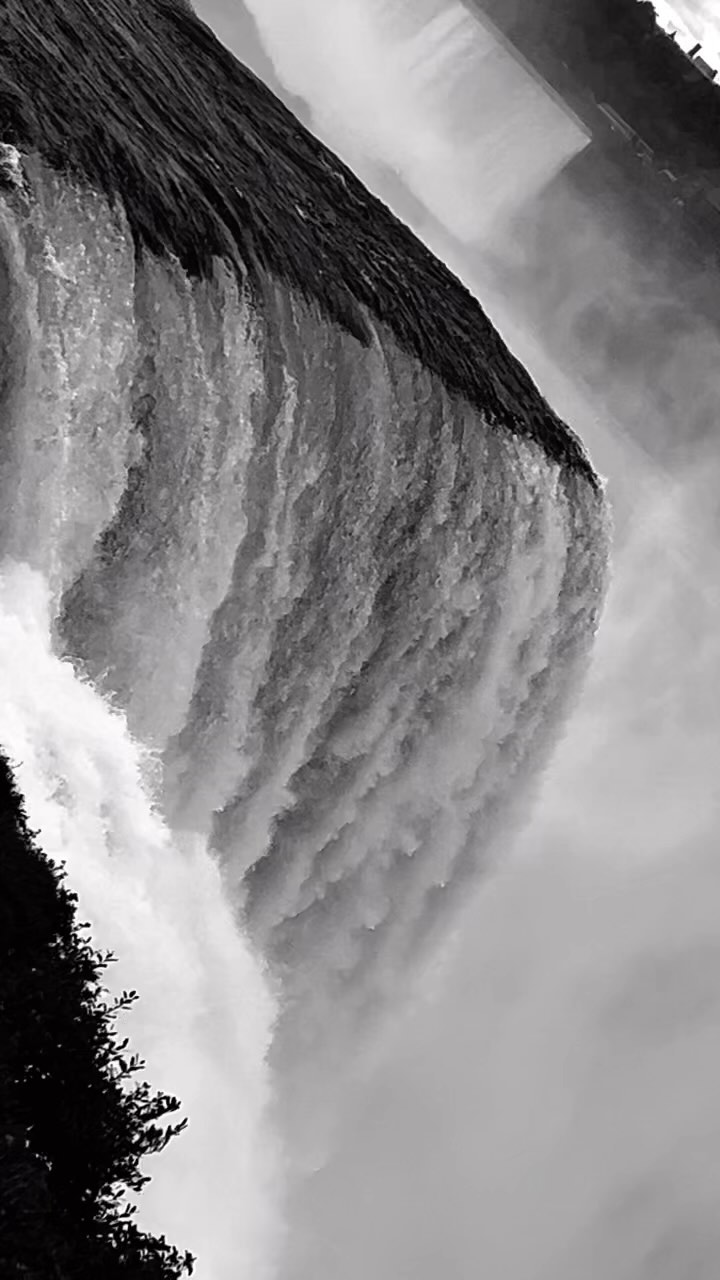 Monochrome photograph captures the cascading rush of a waterfall, its waters churning into mist, set against a backdrop of lush trees.