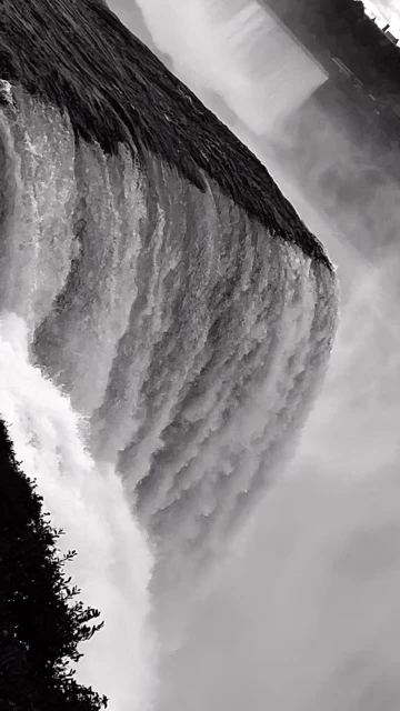 Monochrome photograph captures the cascading rush of a waterfall, its waters churning into mist, set against a backdrop of lush trees.
