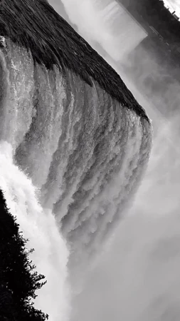 Monochrome photograph captures the cascading rush of a waterfall, its waters churning into mist, set against a backdrop of lush trees.