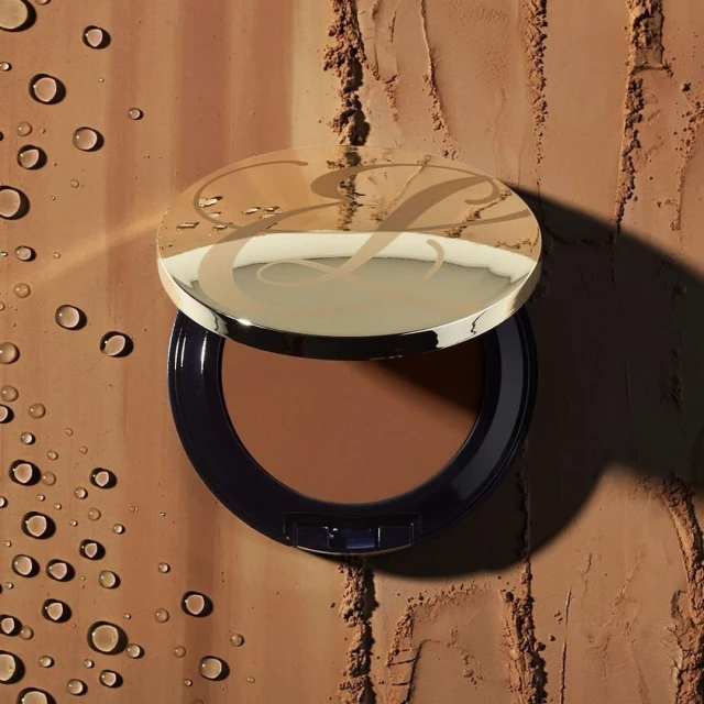 Bronzer makeup powder compact with a shiny gold lid on a brown surface with water droplets and powder textures, beauty and glamour shot.