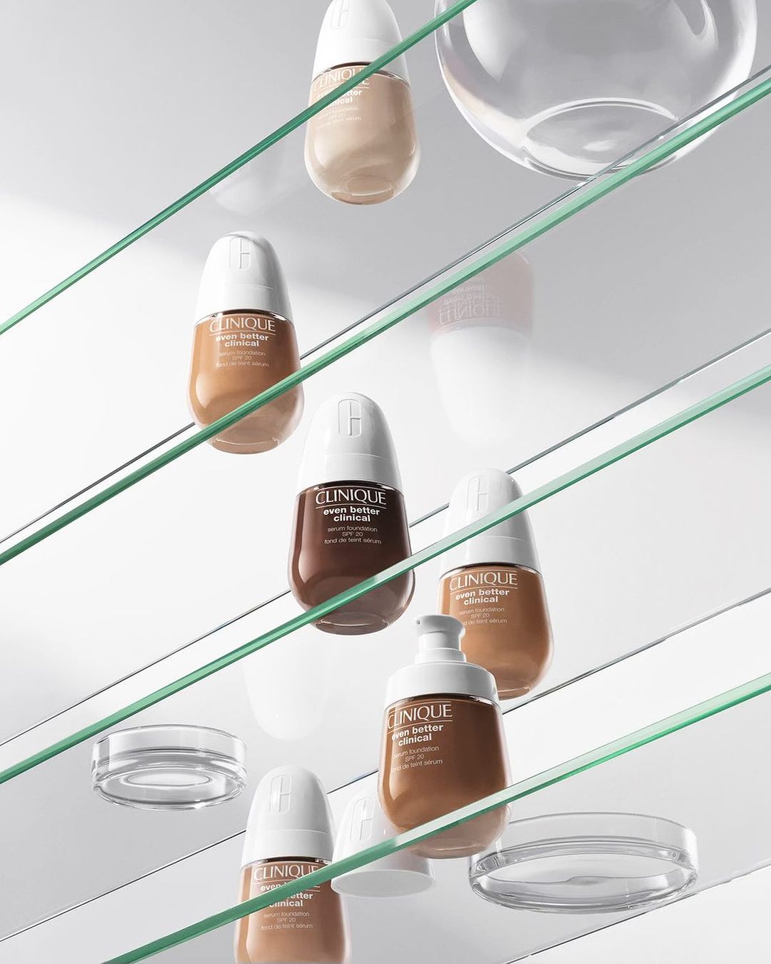 Several Clinique foundation bottles sit on glass shelves with clear bowls in a studio setting, showcasing product assortment.