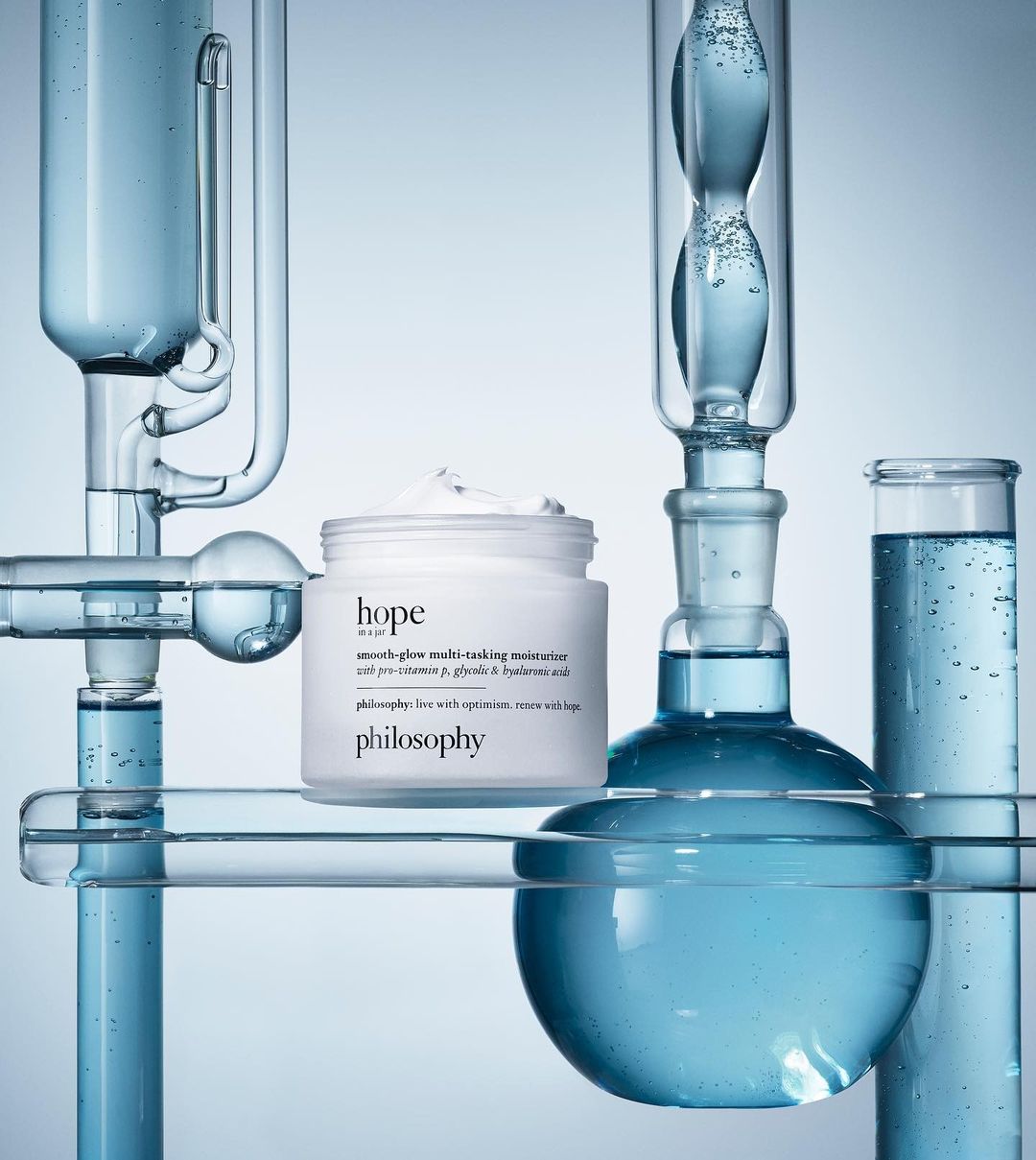 Cosmetic skincare product by Philosophy, 'Hope In A Jar', displayed with lab glassware filled with clear blue liquid on light gradient backdrop.