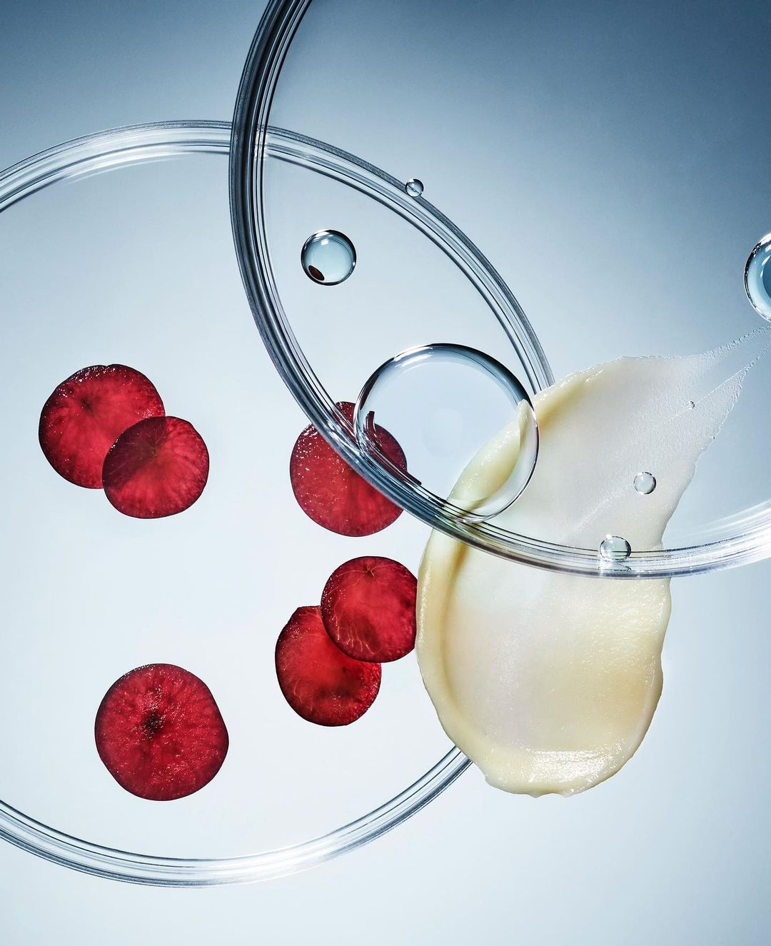 Close-up of cosmetic cream, red fruit slices, and water droplets on clear glass surfaces against a soft blue gradient background.
