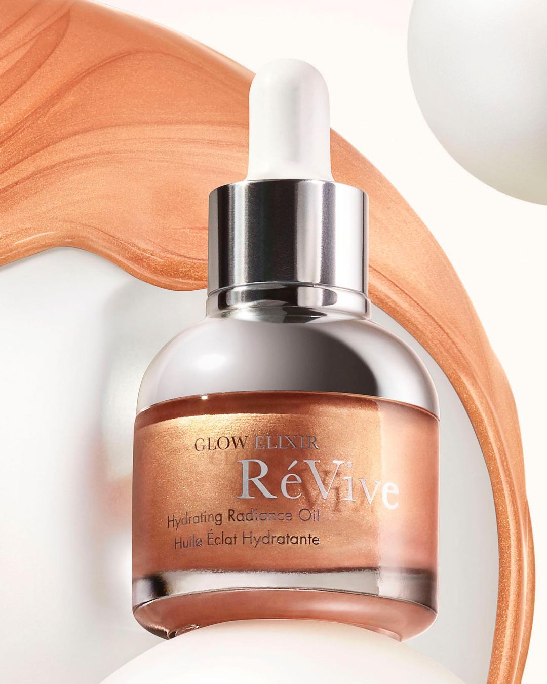 Close-up of a bronze-toned ReVive Glow Elixir Hydrating Radiance Oil bottle with a dropper against a white background with bronze paint streaks.