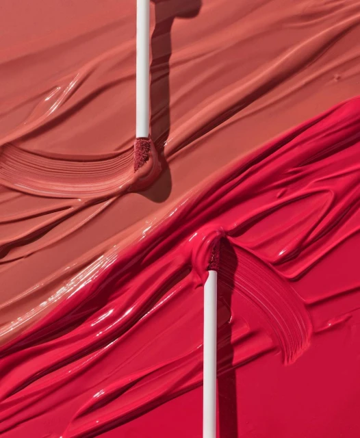 Close-up of makeup product with wand applicator, featuring smears of vibrant red and nude lipstick for a beauty or cosmetic advertisement.