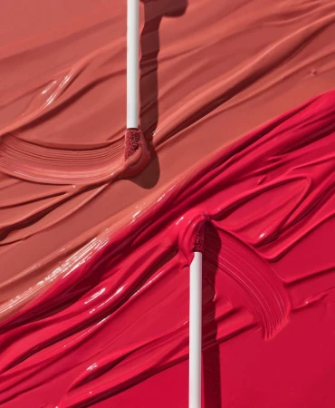 Close-up of makeup product with wand applicator, featuring smears of vibrant red and nude lipstick for a beauty or cosmetic advertisement.