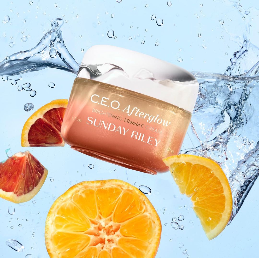 Bright C.E.O. Afterglow brightening Vitamin C cream by Sunday Riley with orange slices and water splashes against light blue background.