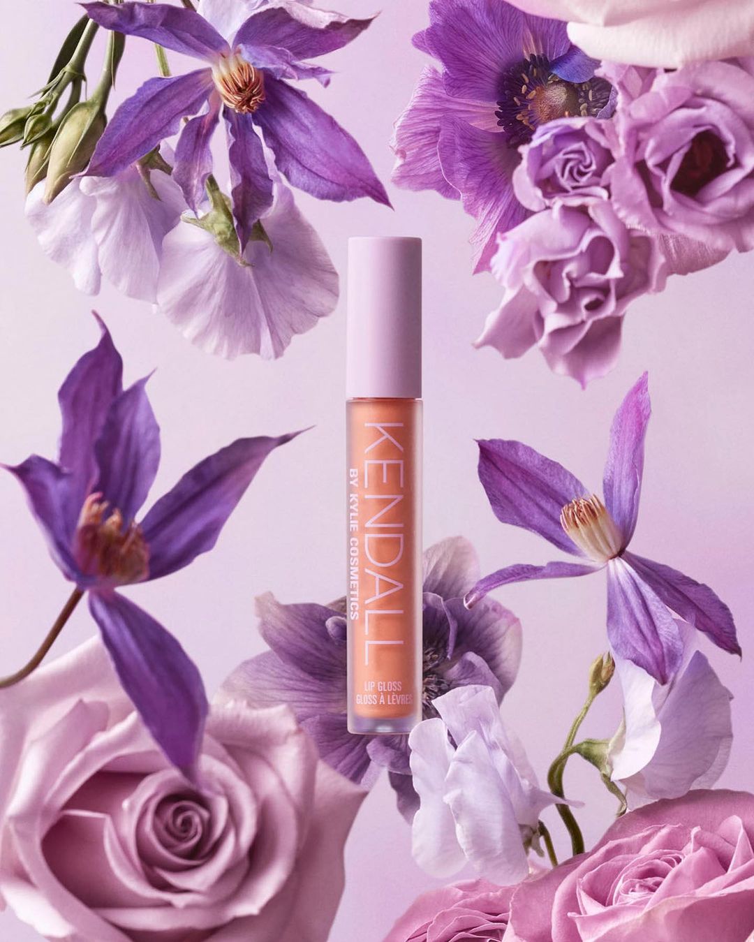 KENDALL by Kylie Cosmetics lip gloss product shot surrounded by lavender, purple, and white roses and other floral accents.