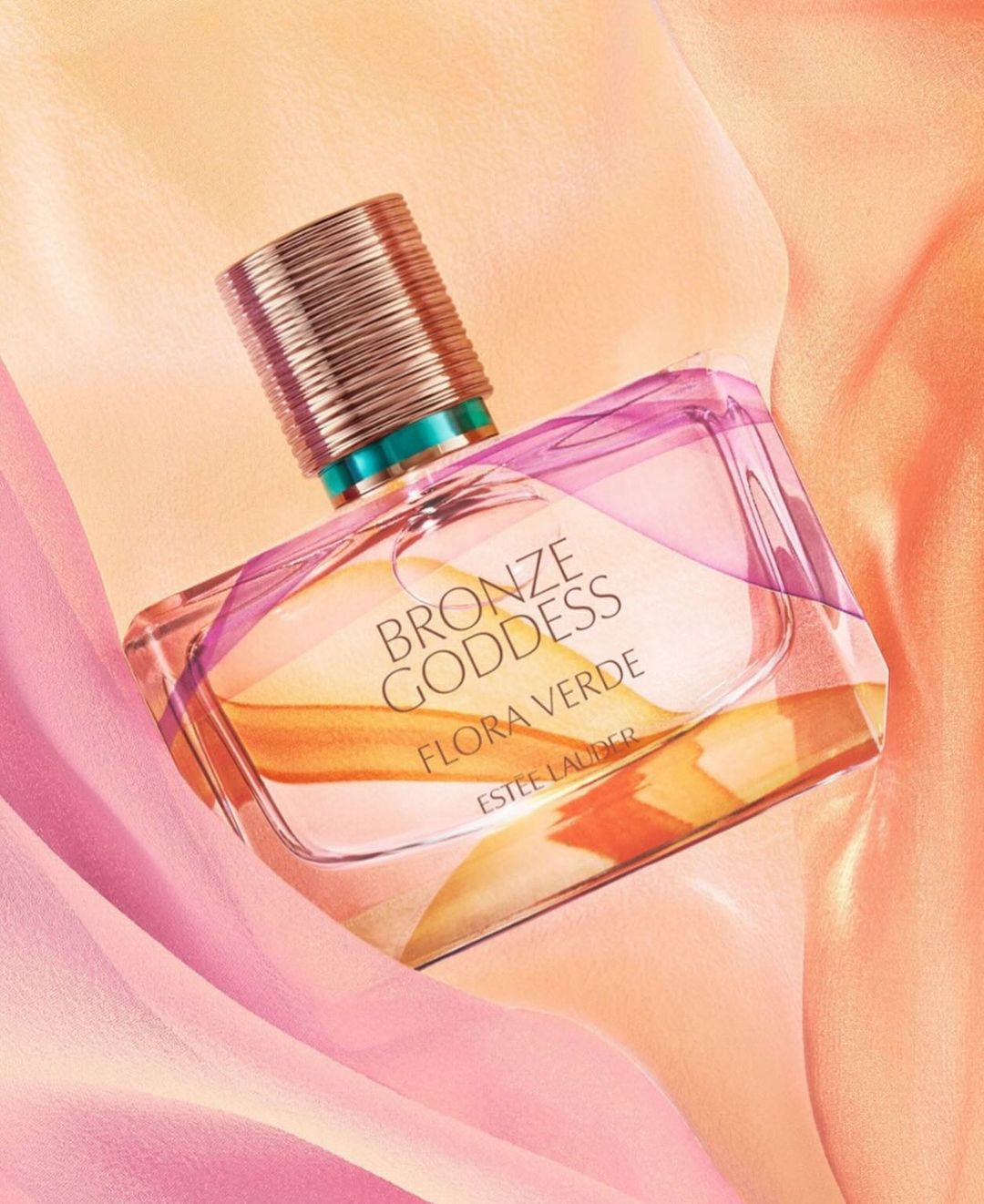 Bronze Goddess Flora Verde perfume bottle by Estee Lauder on a draped peach and pink fabric background in a beauty product shot.