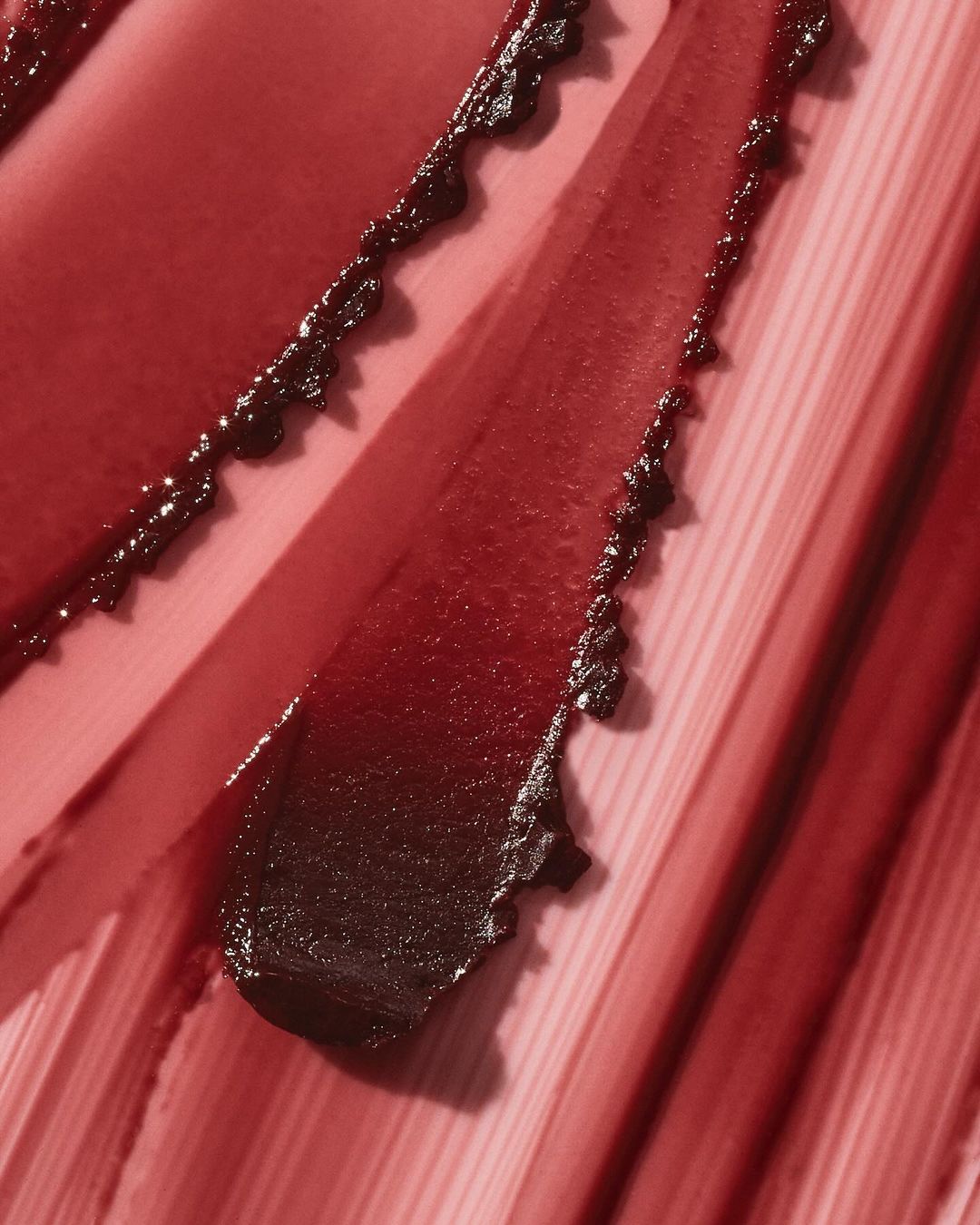 Macro shot of thick, dark red smears of lip gloss. The smears are diagonal and textural, appearing glossy and rich in color.