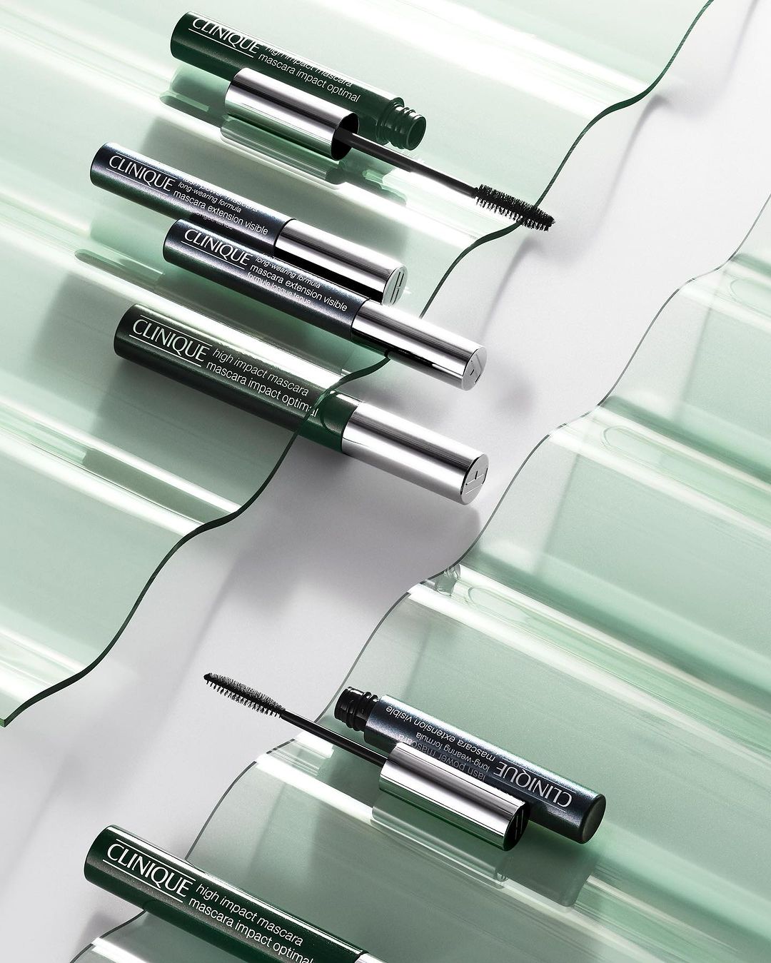 Close-up product shot of Clinique High Impact Mascara tubes in green and silver on a clear, wavy surface creating a fresh, clean beauty aesthetic.