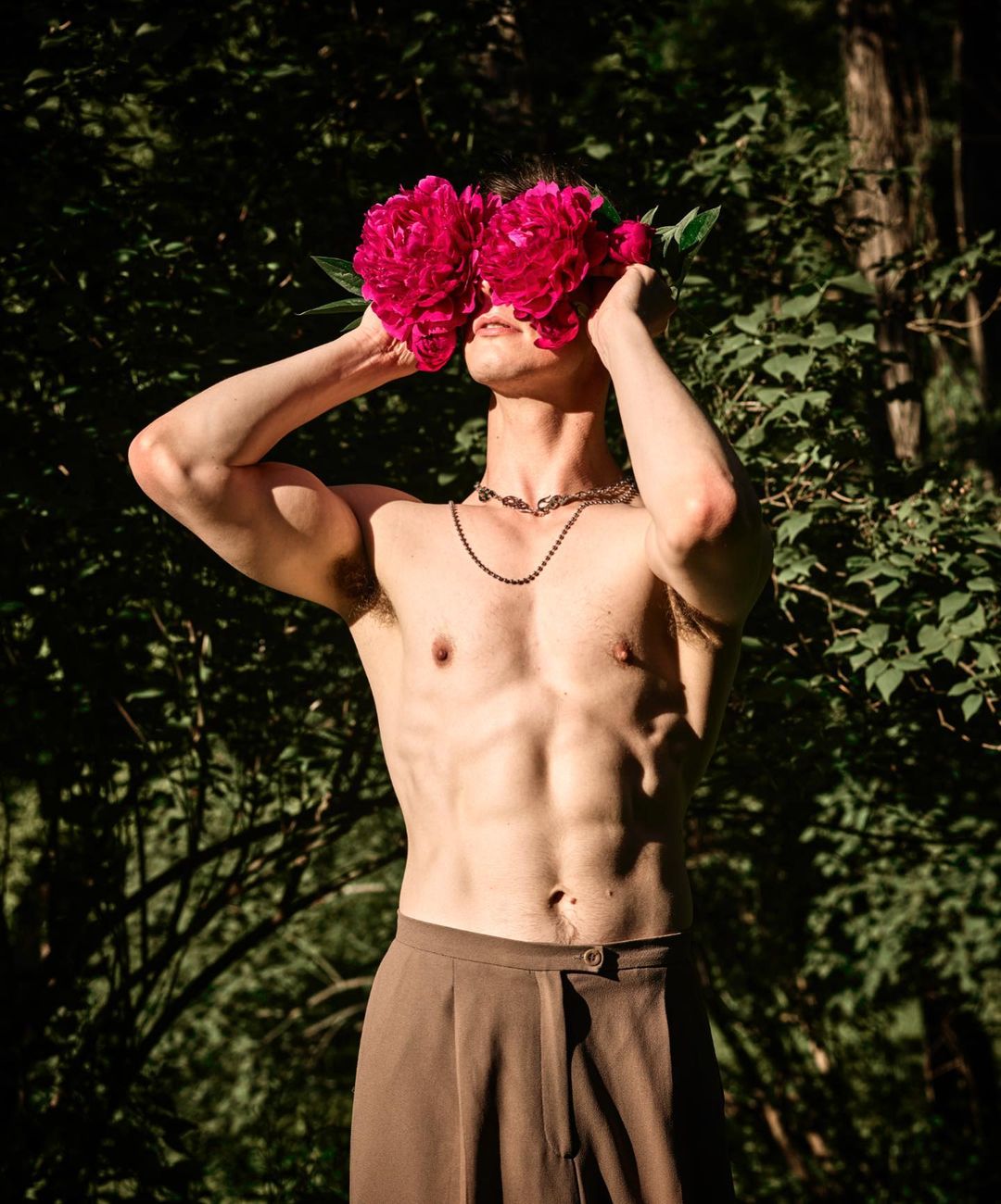 Shirtless male model with dark hair covering face with pink peonies, wearing brown trousers, outdoors in a lush green environment.