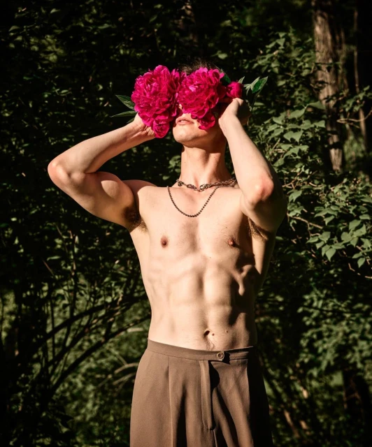 Shirtless male model with dark hair covering face with pink peonies, wearing brown trousers, outdoors in a lush green environment.