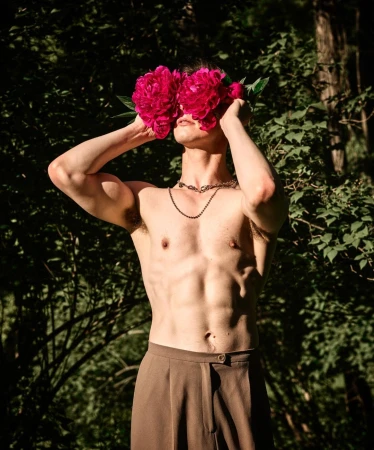 Shirtless male model with dark hair covering face with pink peonies, wearing brown trousers, outdoors in a lush green environment.
