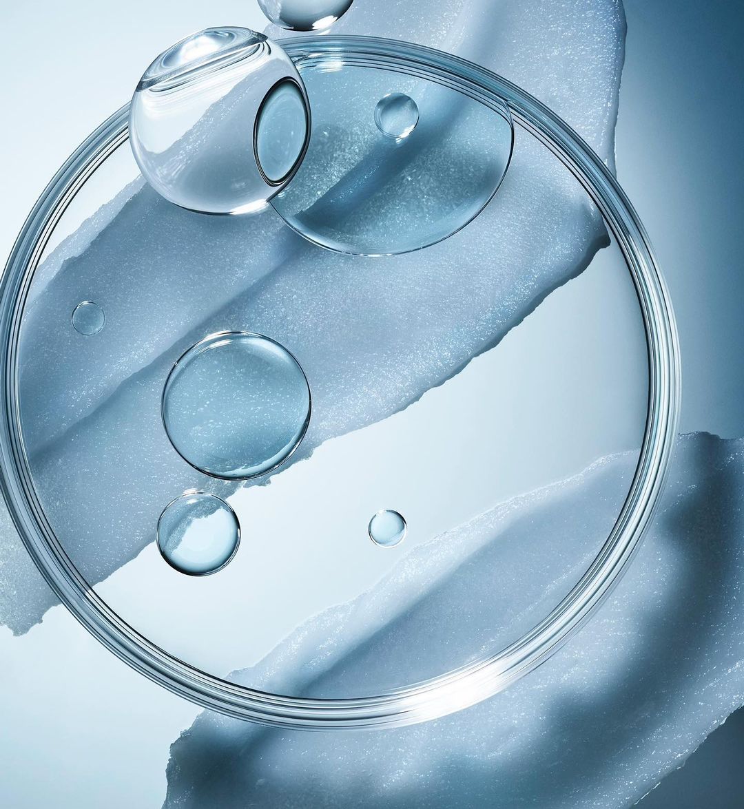 Close-up of a transparent petri dish containing clear liquid, along with water droplets and an irregular texture, set against a blue gradient backdrop.