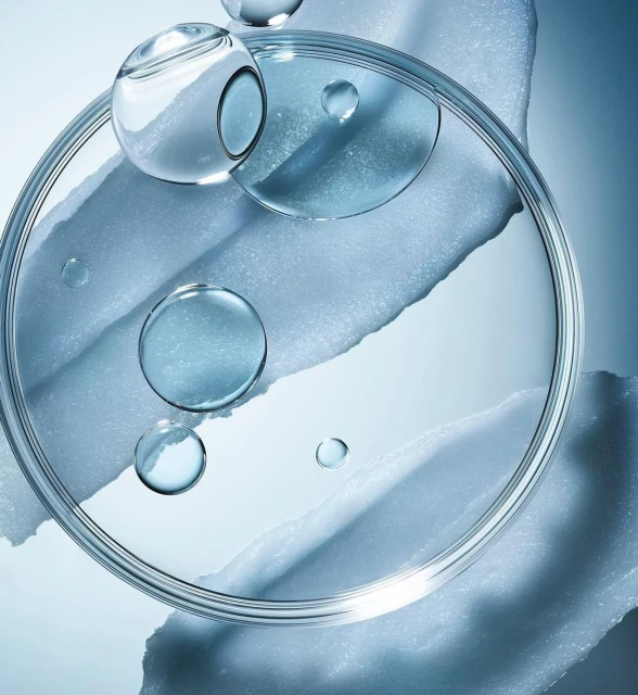 Close-up of a transparent petri dish containing clear liquid, along with water droplets and an irregular texture, set against a blue gradient backdrop.