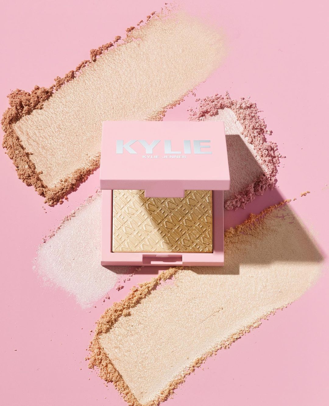 Kylie Cosmetics powder compact with light gold and pink powder makeup in a square pink case on a pink backdrop with loose makeup.