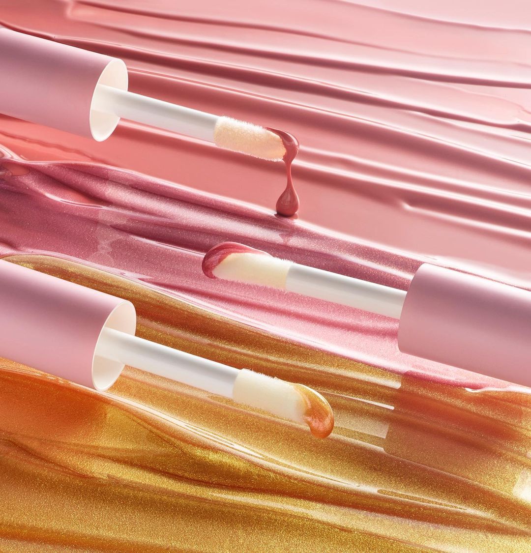 Macro studio shot of lip gloss applicators with pink and gold product on a glossy surface with matching color streaks.