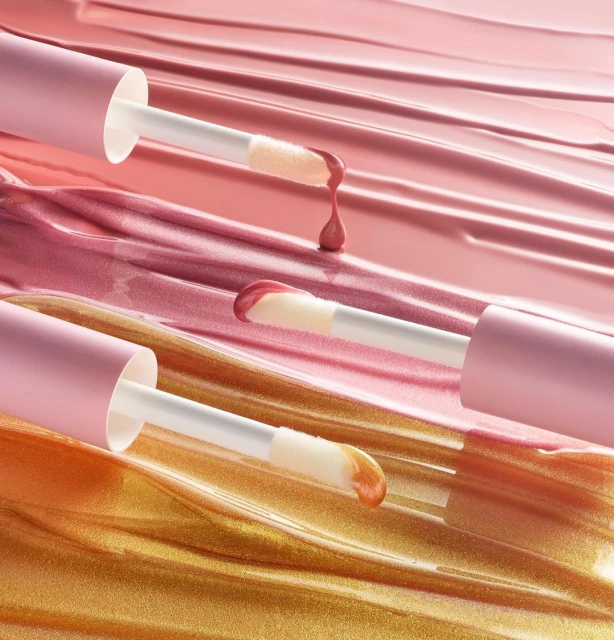 Macro studio shot of lip gloss applicators with pink and gold product on a glossy surface with matching color streaks.