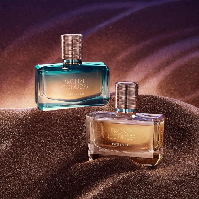 Two 'Bronze Goddess' perfume bottles by Estee Lauder on a textured bronze surface, set against a purple-to-gold gradient background.