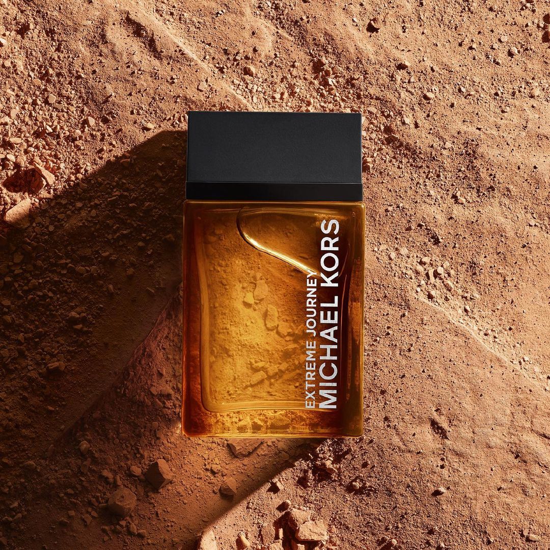 Close-up of Michael Kors 'Extreme Journey' fragrance bottle set on a textured brown surface, accentuating its amber color and design.