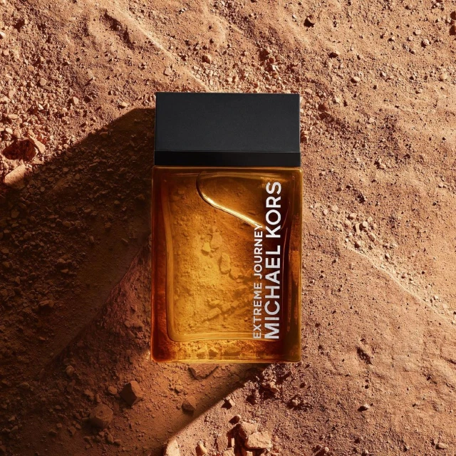 Close-up of Michael Kors 'Extreme Journey' fragrance bottle set on a textured brown surface, accentuating its amber color and design.