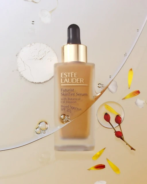Estee Lauder Futurist SkinTint Serum in a square bottle with a dropper, surrounded by petals and powder on a light background.