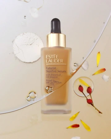 Estee Lauder Futurist SkinTint Serum in a square bottle with a dropper, surrounded by petals and powder on a light background.