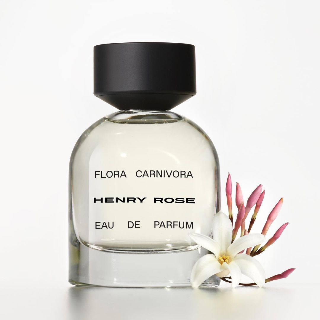 Close-up product shot of a Henry Rose Flora Carnivora perfume bottle with a black cap, set against a white background with a flower.