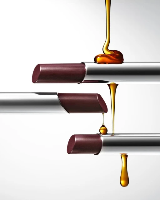 Three plum lipsticks arranged in a row with golden liquid dripping over them on a stark white backdrop for a beauty advertisement.