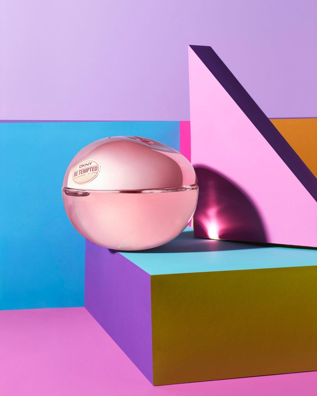 Close-up of DKNY Be Tempted perfume bottle on a geometric platform with purple, pink, teal, and yellow geometric shapes behind it.