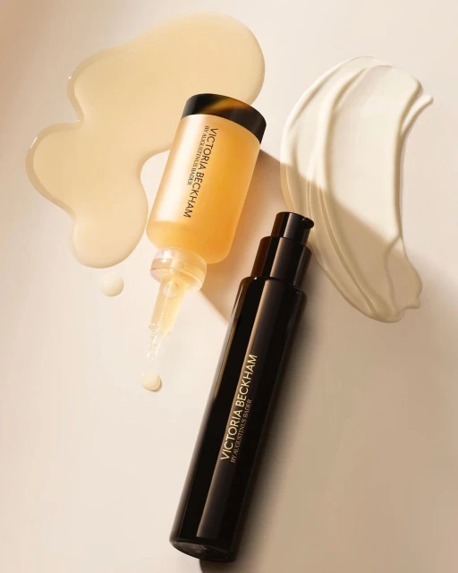 Close-up still life of Victoria Beckham skincare products with serum, moisturizer, and oil textures on a neutral-toned background.